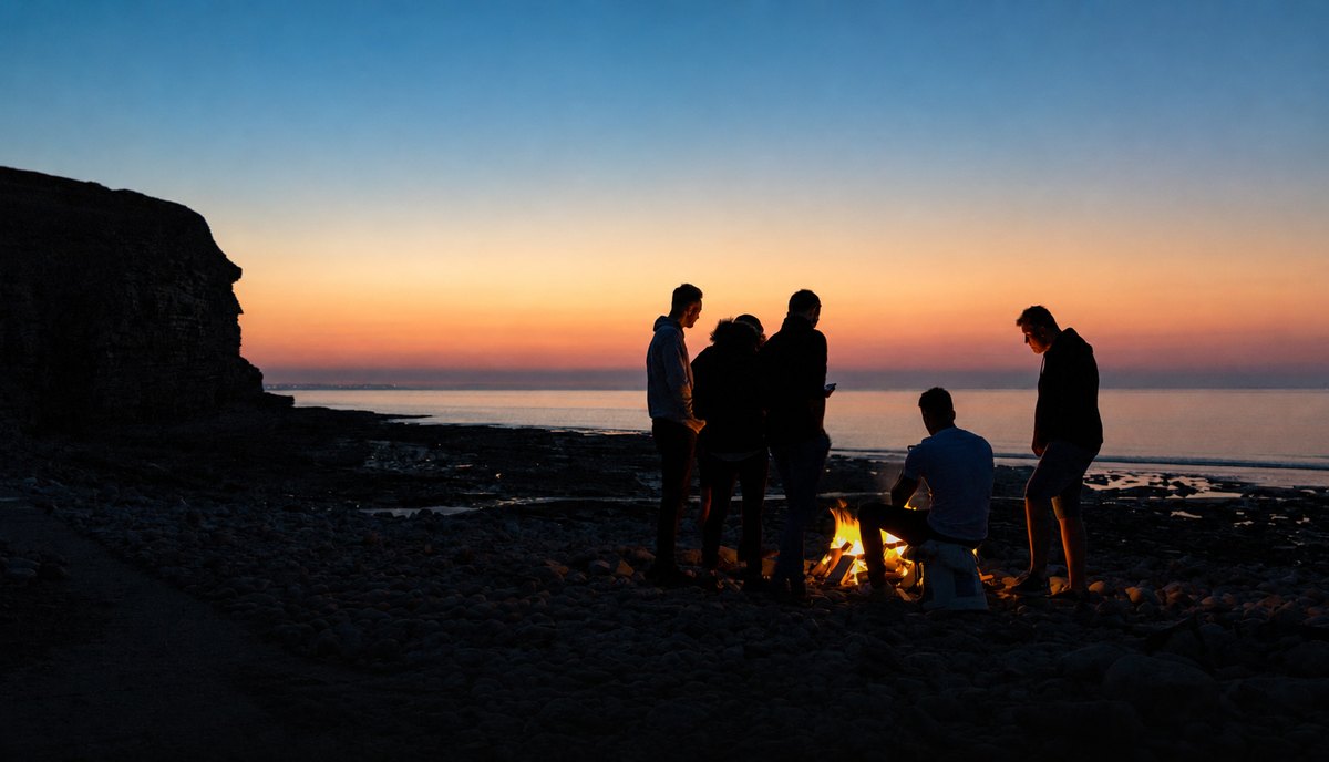 A fire circle at dusk on the shore, the sea and last light behind